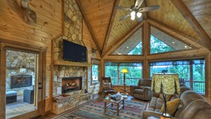 A spacious living room with a stone fireplace, wooden ceiling, and large windows offering a view of the outdoors. - Deer Trails Cabin - Blue Ridge (Cherry Log)