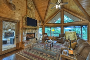 A spacious living room with a stone fireplace, wooden ceiling, and large windows offering a view of the outdoors.
