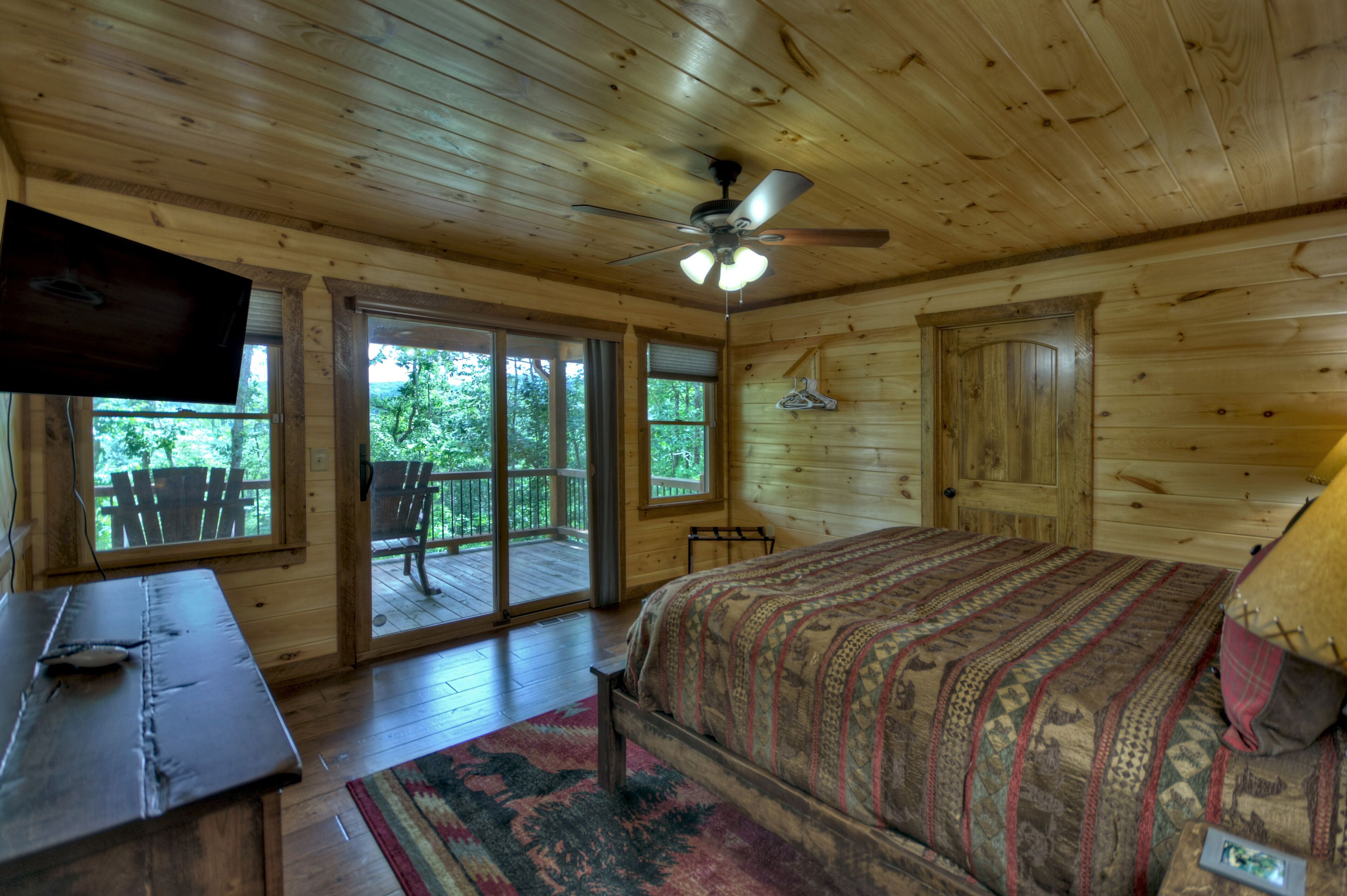 A wooden cabin bedroom with a bed, a desk, a television, and a ceiling fan.