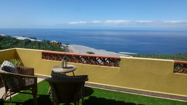 A balcony with wicker chairs and a small table, overlooking a coastal view with the ocean and a distant shoreline. - Apartment Rosa 1 - Apartments Rosa (Tijarafe)