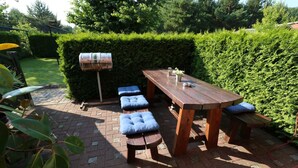 Une terrasse avec une table et des bancs en bois, un barbecue et une haie en arrière-plan.