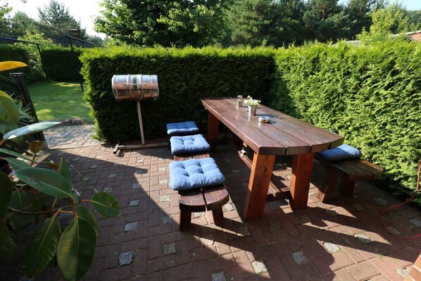 A patio with a wooden table and benches, a barbecue grill, and a hedge in the background.