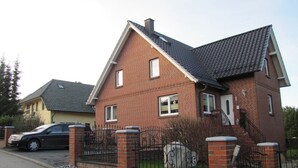 Une maison à deux étages, construite en briques, avec un toit en tuiles, une allée bordée d’une clôture et une voiture garée.