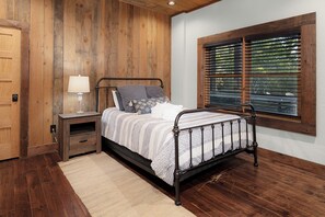 Quarto com parede revestida de madeira, cama com estrutura de metal, mesa de cabeceira com abajur e janela com persianas.