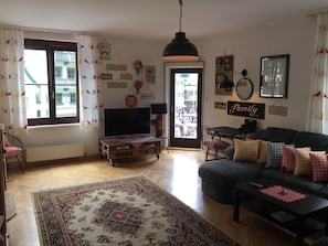 Ett vardagsrum med en tv, en soffa och ett soffbord. Det finns en dörr som leder ut, och rummet har en matta och dekorationer på väggarna.