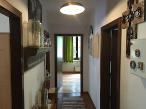 Un couloir avec un plancher en bois, un luminaire suspendu, une porte sur la droite et, au fond, une fenêtre avec des rideaux verts.