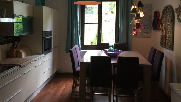 A kitchen with a dining table, chairs, a hanging light fixture, and a window with curtains. - Ferienwohnung 3 - Ferienwohnungen in der Kaffeewerkstatt (Sankt Wolfgang im Salzkammergut)