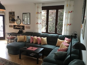Un salon comprenant un canapé d’angle, une table basse, une table à manger avec des chaises et une grande fenêtre avec des rideaux.