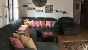 A living room with a large round wall clock, a sofa with multiple pillows, a small table with a remote control, and a floor lamp. - Ferienwohnung 3 - Ferienwohnungen in der Kaffeewerkstatt (Sankt Wolfgang im Salzkammergut)