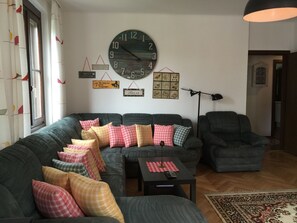 Ett vardagsrum med en stor, rund väggklocka, en soffa med många kuddar, ett litet bord med en fjärrkontroll och en golvlampa.