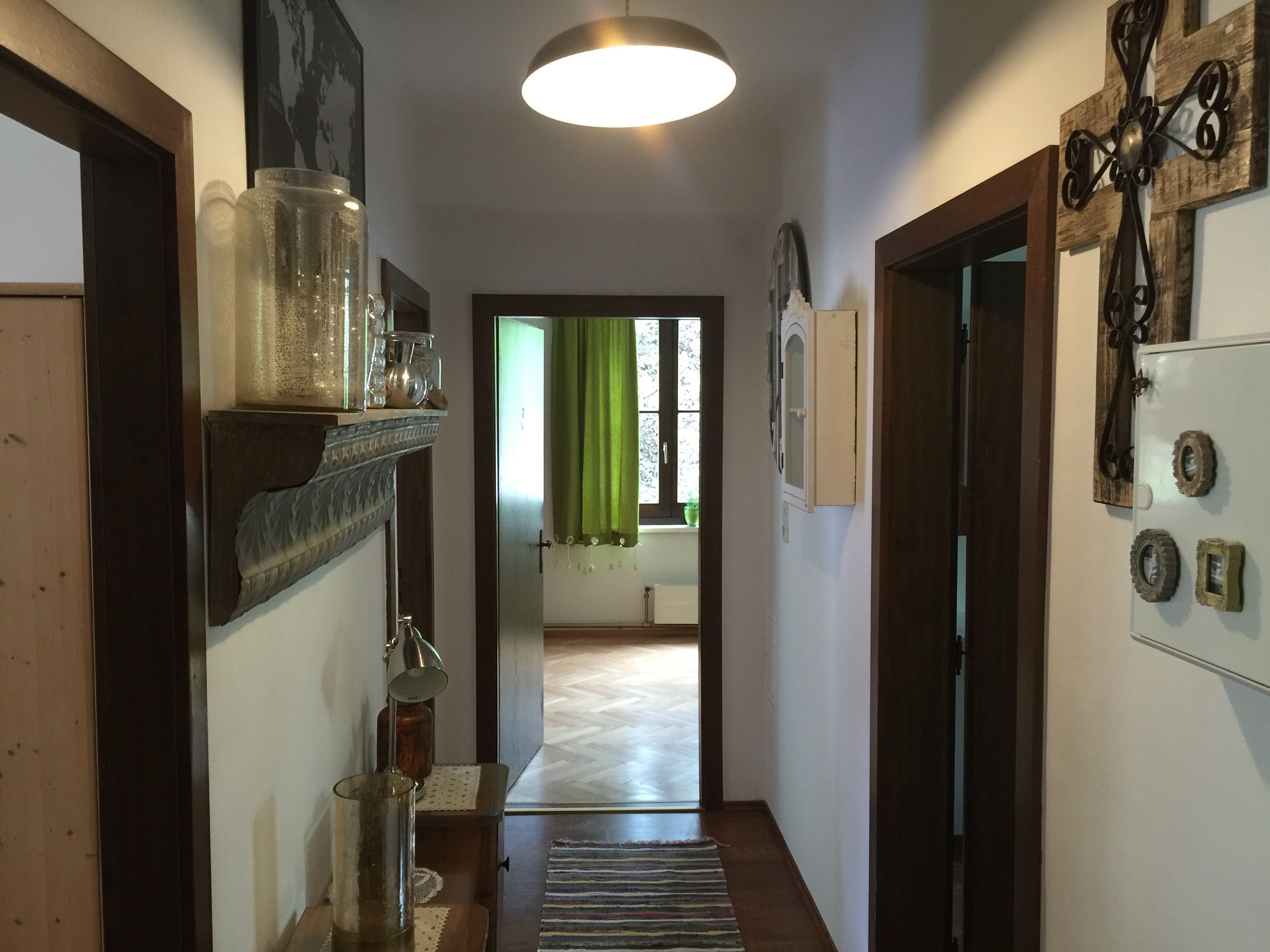 Un couloir avec un plancher en bois, un luminaire suspendu, une porte sur la droite et, au fond, une fenêtre avec des rideaux verts.