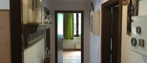 Un couloir avec un plancher en bois, un luminaire suspendu, une porte sur la droite et, au fond, une fenêtre avec des rideaux verts.