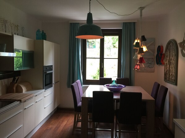 A kitchen with a dining table, chairs, a hanging light fixture, and a window with curtains.