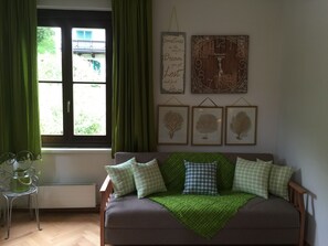 A room with a window, a sofa with green and plaid cushions, a wall clock, and framed botanical prints.