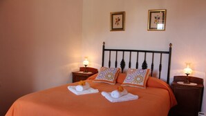 Ein ordentlich gemachtes Bett mit orangefarbener Tagesdecke, zwei Zierkissen und zwei mit Blumen verzierten Handtüchern. Zwei Nachttische mit Lampen stehen bereit, und das Zimmer hat ein schlichtes, sauberes Design.