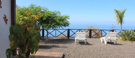 A patio with two white lounge chairs, a potted plant, and a view of the ocean.