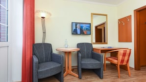 A hotel room with two grey armchairs, a small round table with a bottle, a television on the wall, and a wooden door.