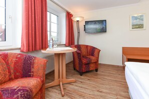 Et værelse med en stribet sofa, et lille bord, en stol, en seng, et vindue med gardiner, et vægmonteret fjernsyn og et indrammet billede.