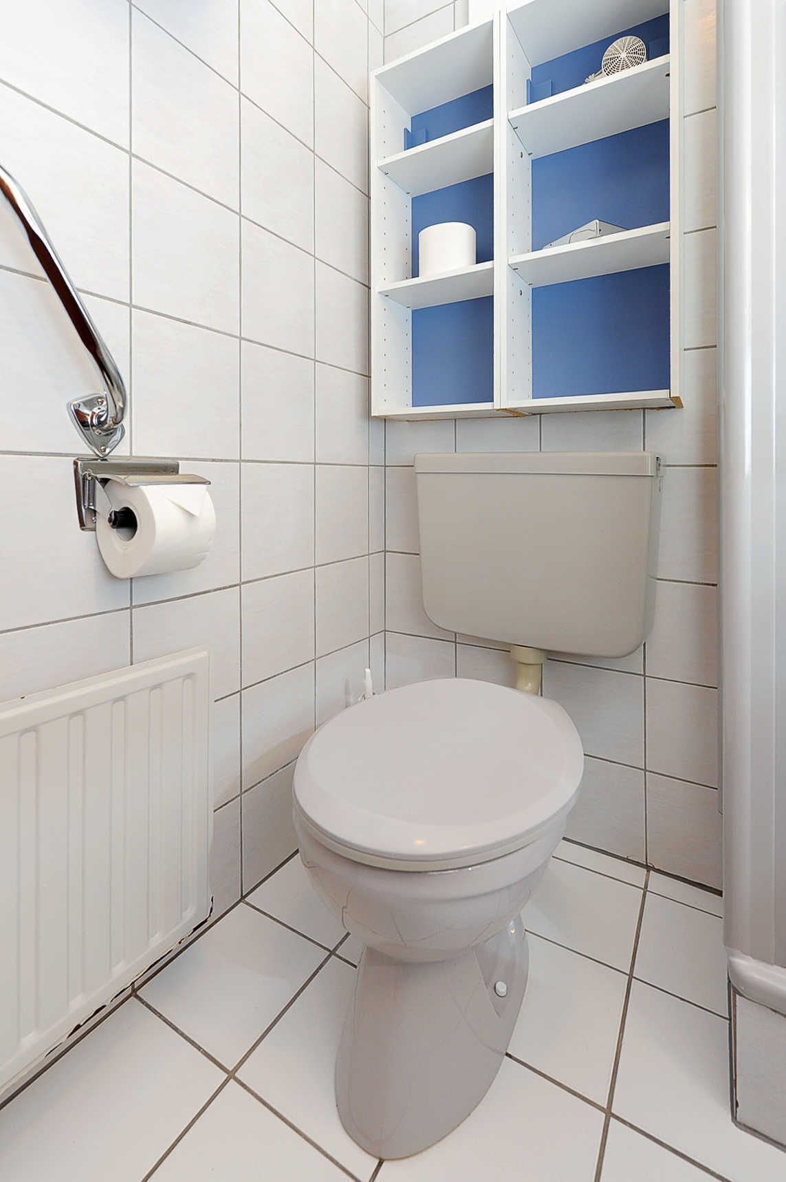 Une salle de bain avec des murs carrelés de blanc, des toilettes blanches, un radiateur blanc et une étagère bleue avec un porte-rouleau de papier toilette.