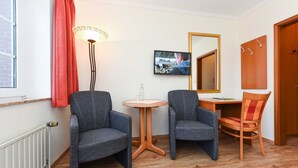 A room with two grey armchairs, a small round table, a floor lamp, a television, a mirror, and a wooden door.