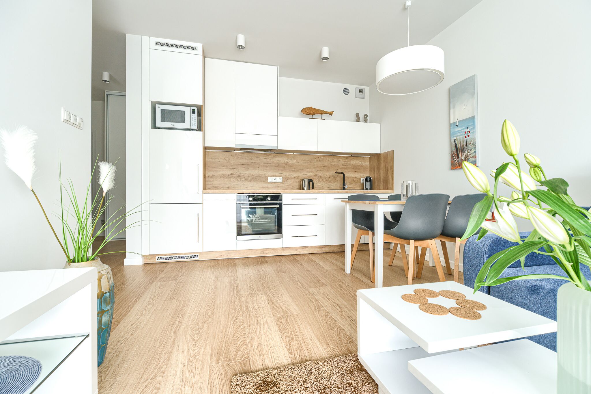 Una cocina moderna con armarios blancos, microondas y horno. Hay un comedor con mesa y sillas, y una sala de estar con sofá y mesa de centro.