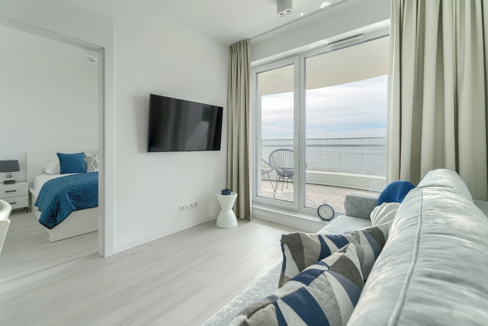 A modern hotel room with a large window, a flat-screen TV, a bed with blue bedding, a white sofa, and a small side table.