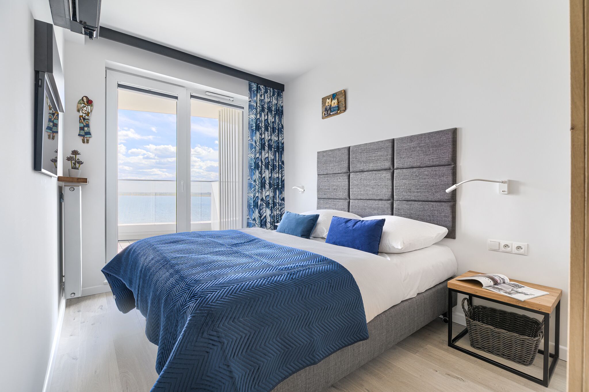 A modern bedroom with a large bed, a grey headboard, a bedside table with a lamp, and a view of the sky through the window.
