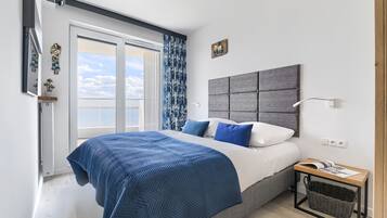 A modern bedroom with a large bed, a grey headboard, a bedside table with a lamp, and a view of the sky through the window.