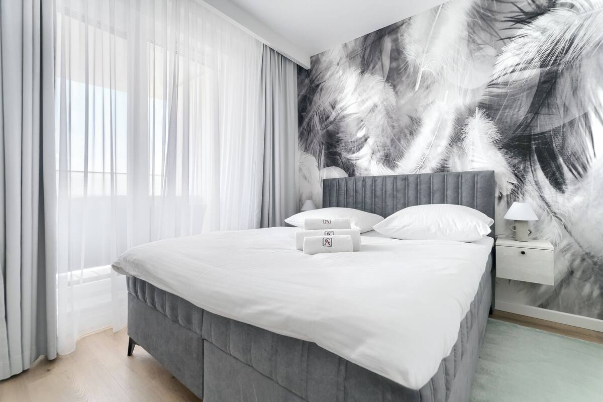 Un dormitori modern amb un llit gris, roba de llit blanca i un mural de plomes a la paret.