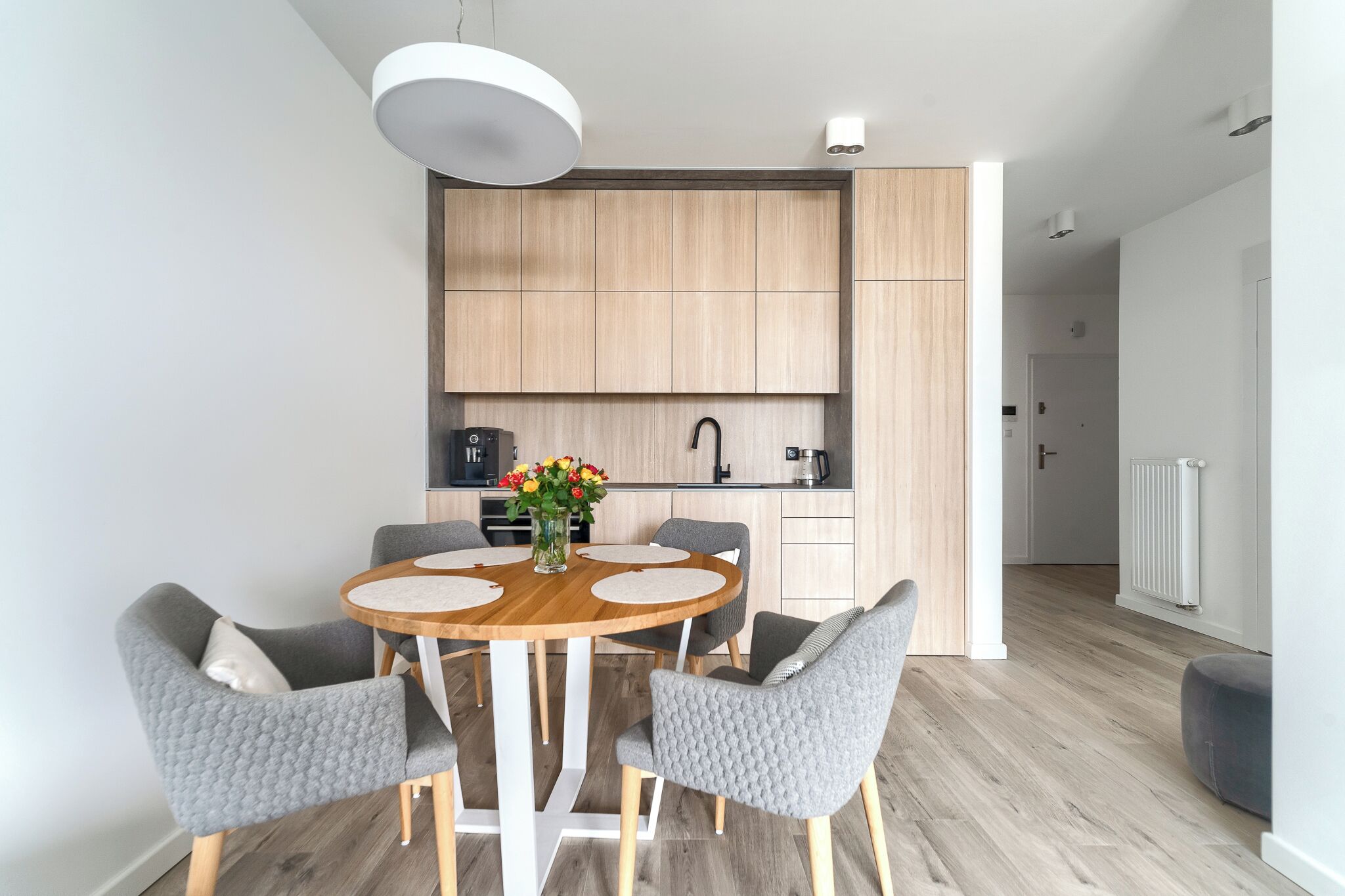 Eine moderne Küche mit Holzschränken, einem runden Esstisch mit grau bezogenen Stühlen und einem Blumenstrauß auf dem Tisch.