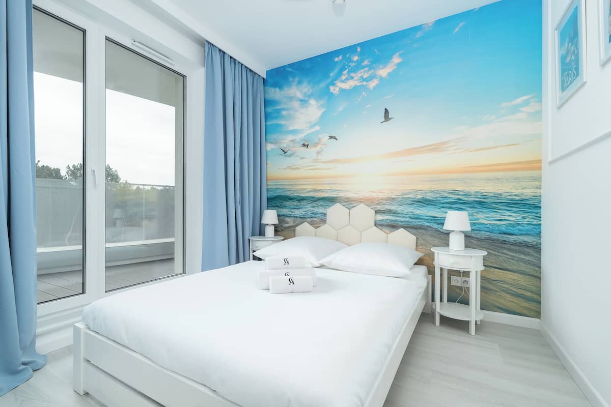 Un dormitori amb una finestra gran, un llit amb roba de llit blanca, un mural d'un tramuntana sobre la mar i dues taules de nit amb làmpades.