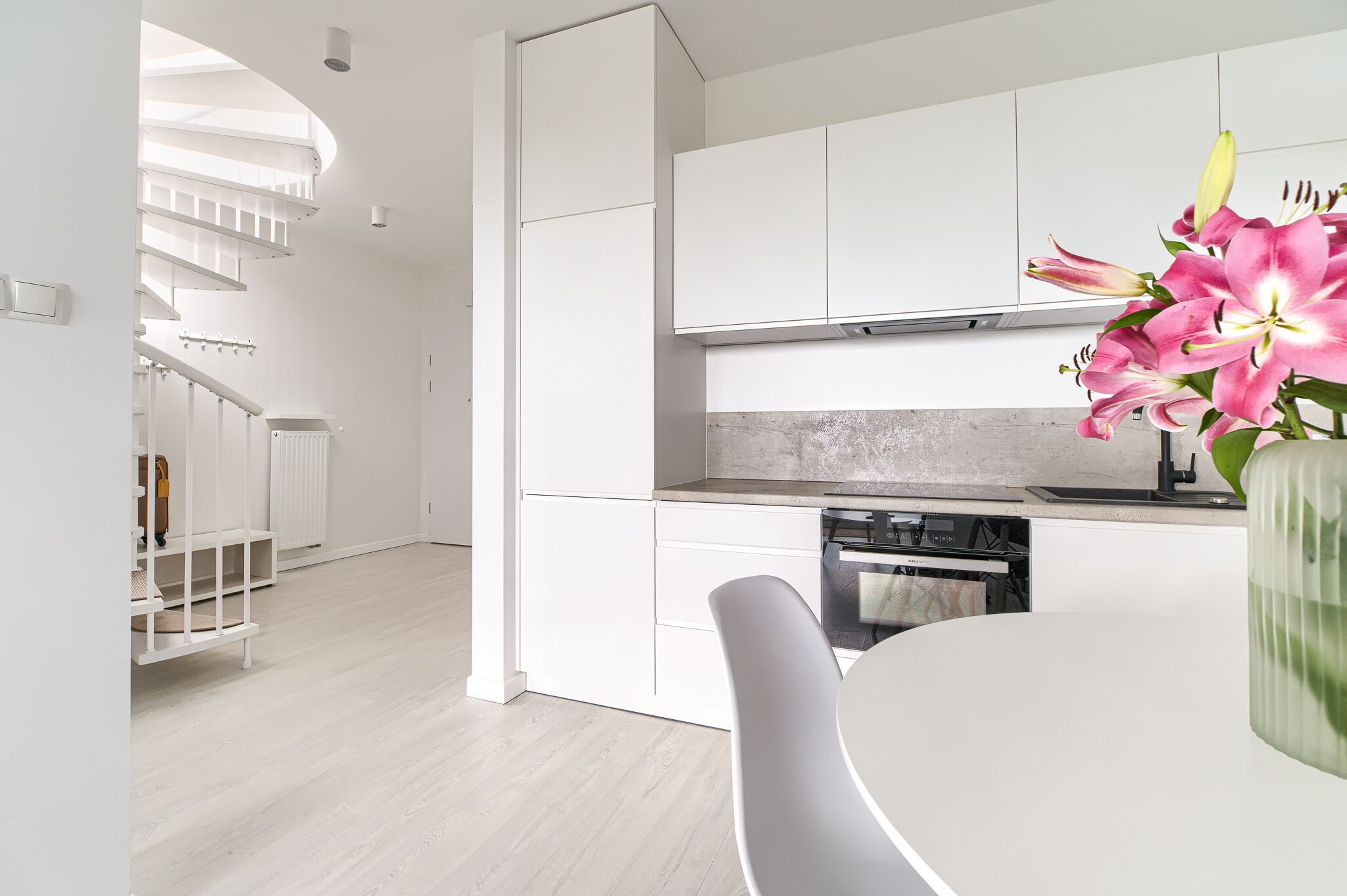 Una cocina moderna con armarios blancos, horno empotrado y comedor con mesa y silla blancos.
