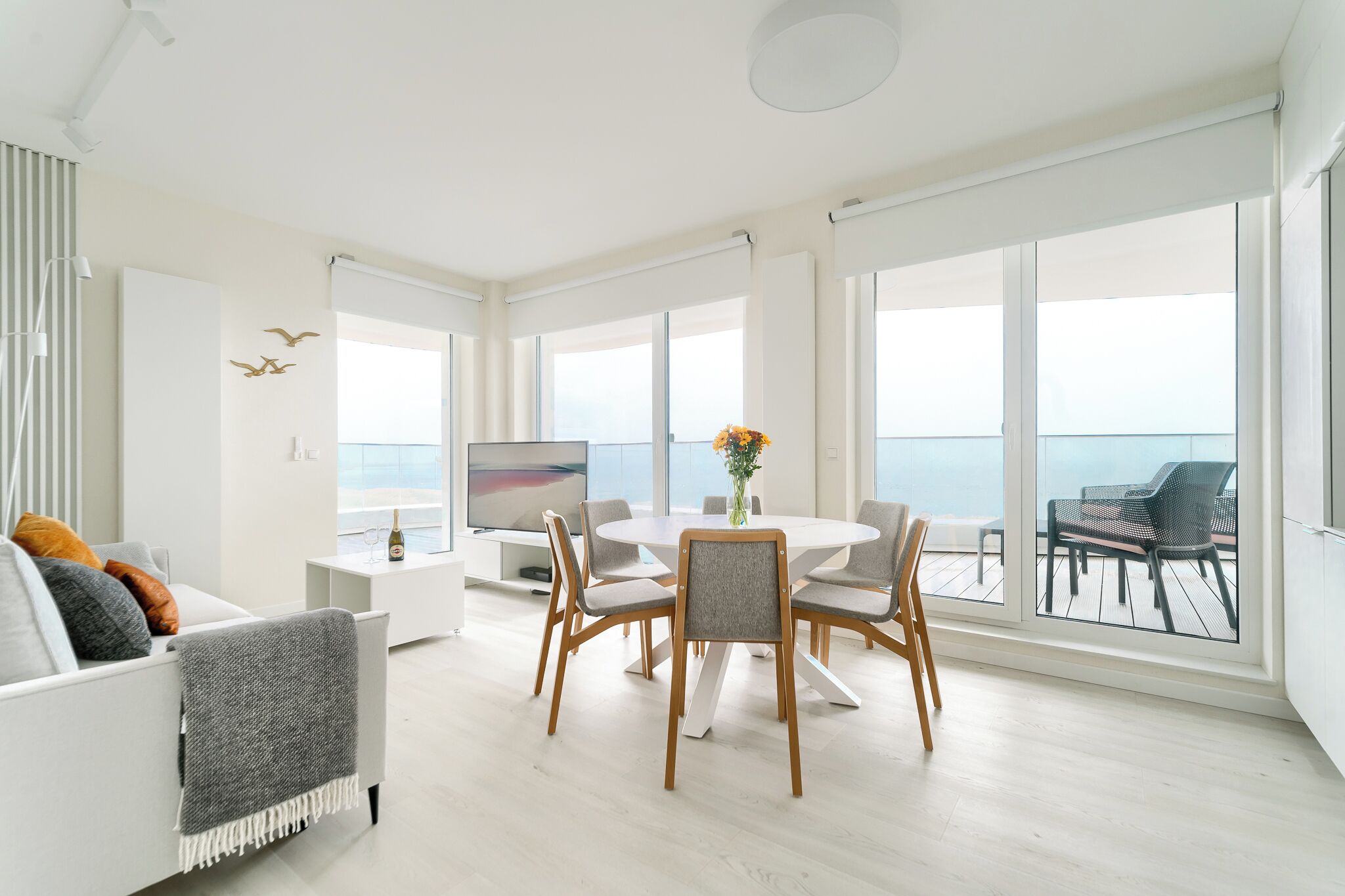 Un soggiorno moderno con un tavolo da pranzo rotondo, posti a sedere confortevoli, una TV a schermo piatto e ampie finestre con vista sul mare.