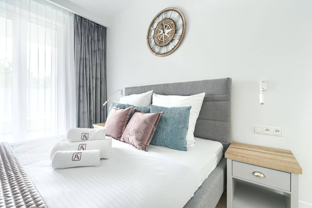 Habitació d'hotel moderna amb un llit gran, una taula de nit, un rellotge de paret i una finestra amb cortines.