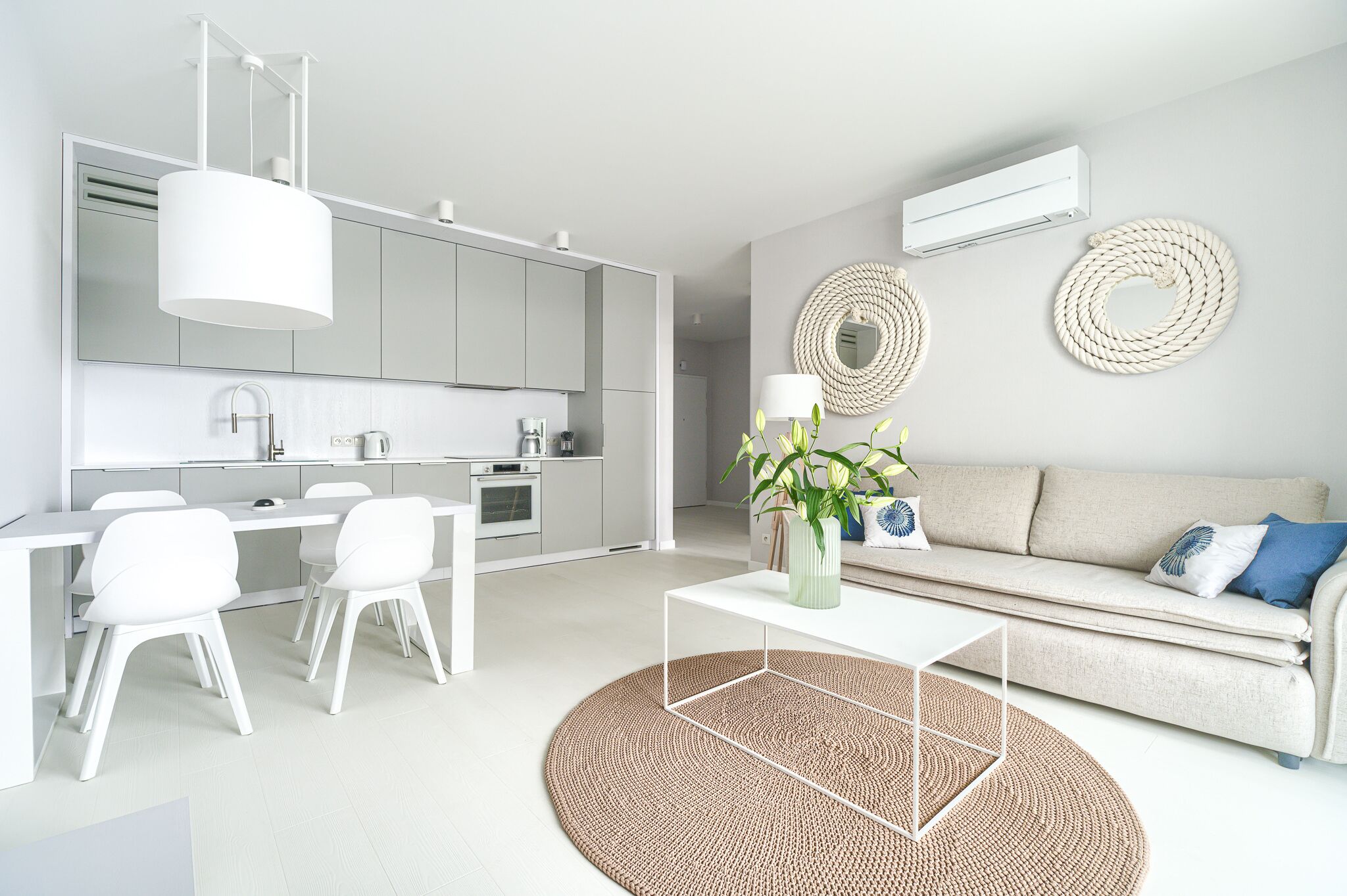 A modern kitchen with white cabinets, a dining area with white chairs, a sofa with blue pillows, and a round woven rug.