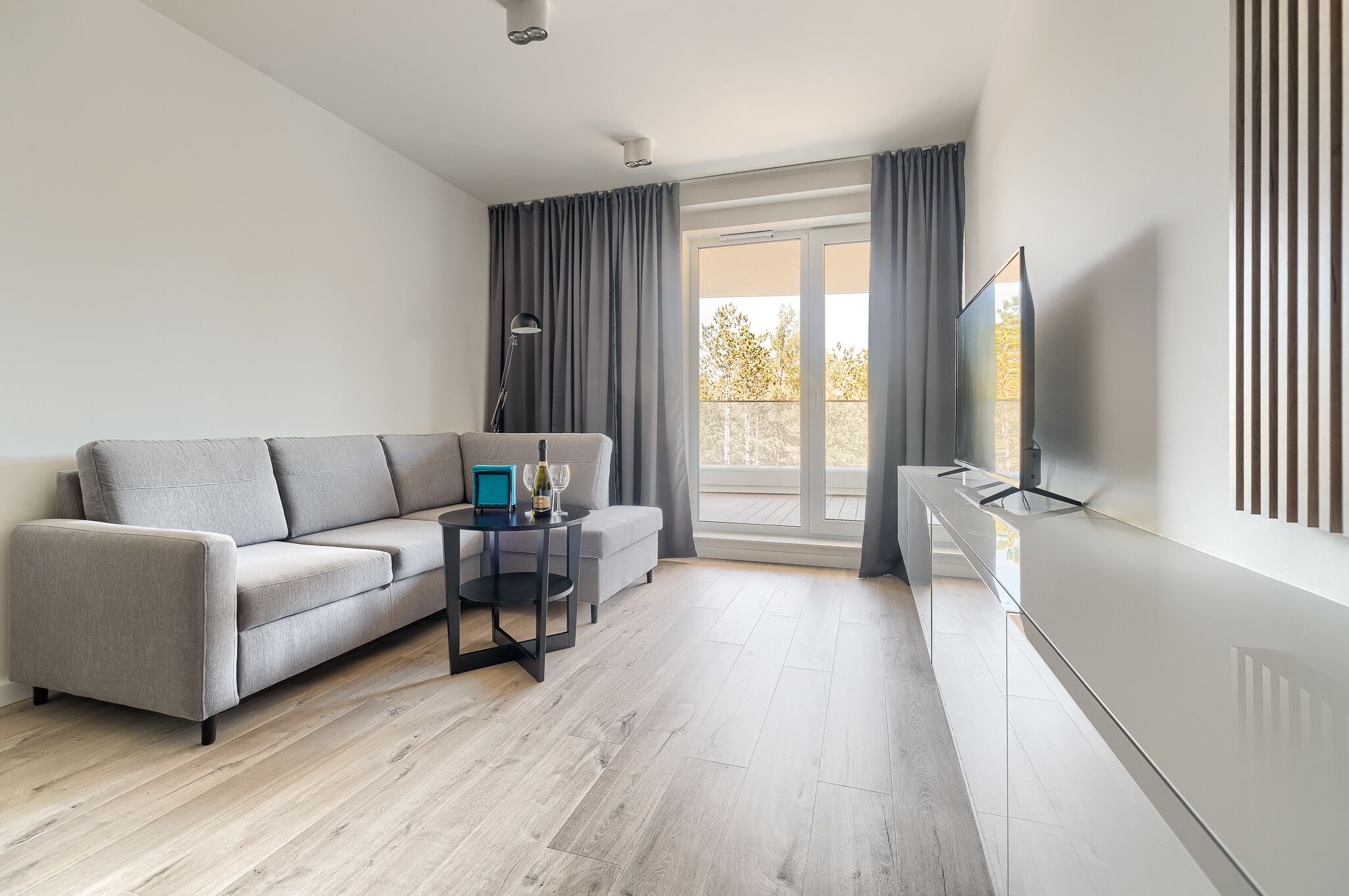 A modern living room with a grey sofa, a small table with a wine bottle, and a TV on a stand.
