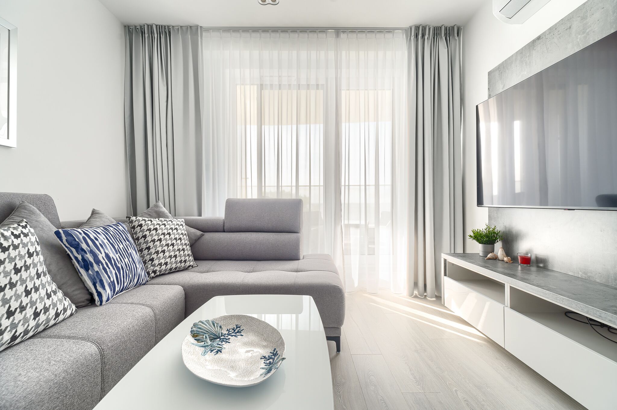 A modern living room with a grey sofa, a white coffee table, and a large flat-screen TV on a white console.