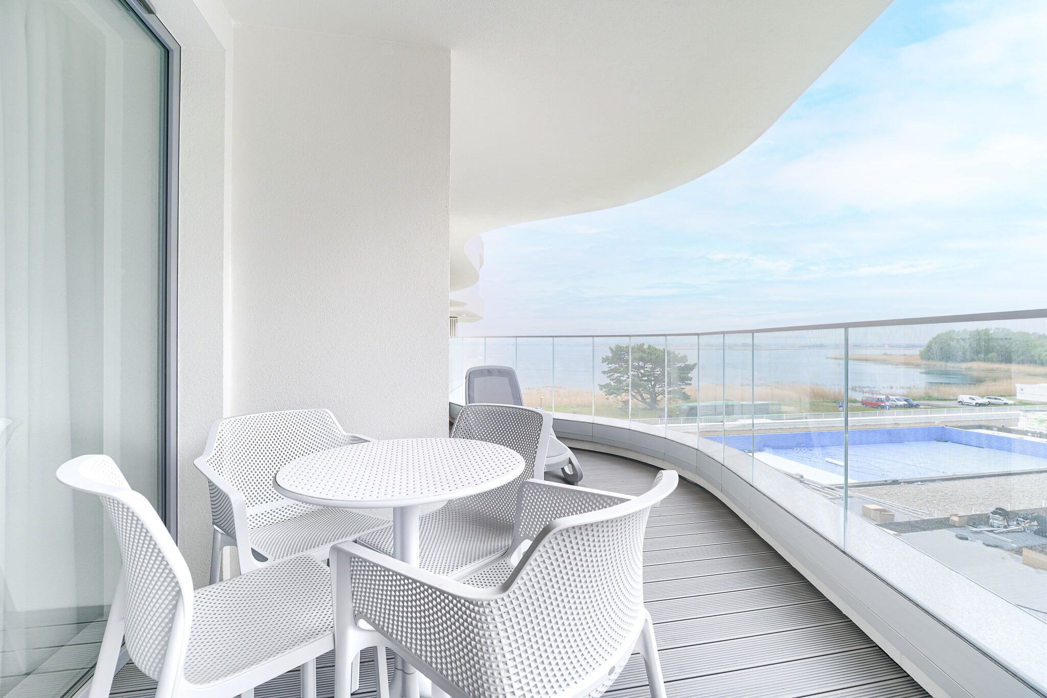 Un balcón con una mesa redonda y sillas blancas, con vistas a una piscina y paisaje distante.