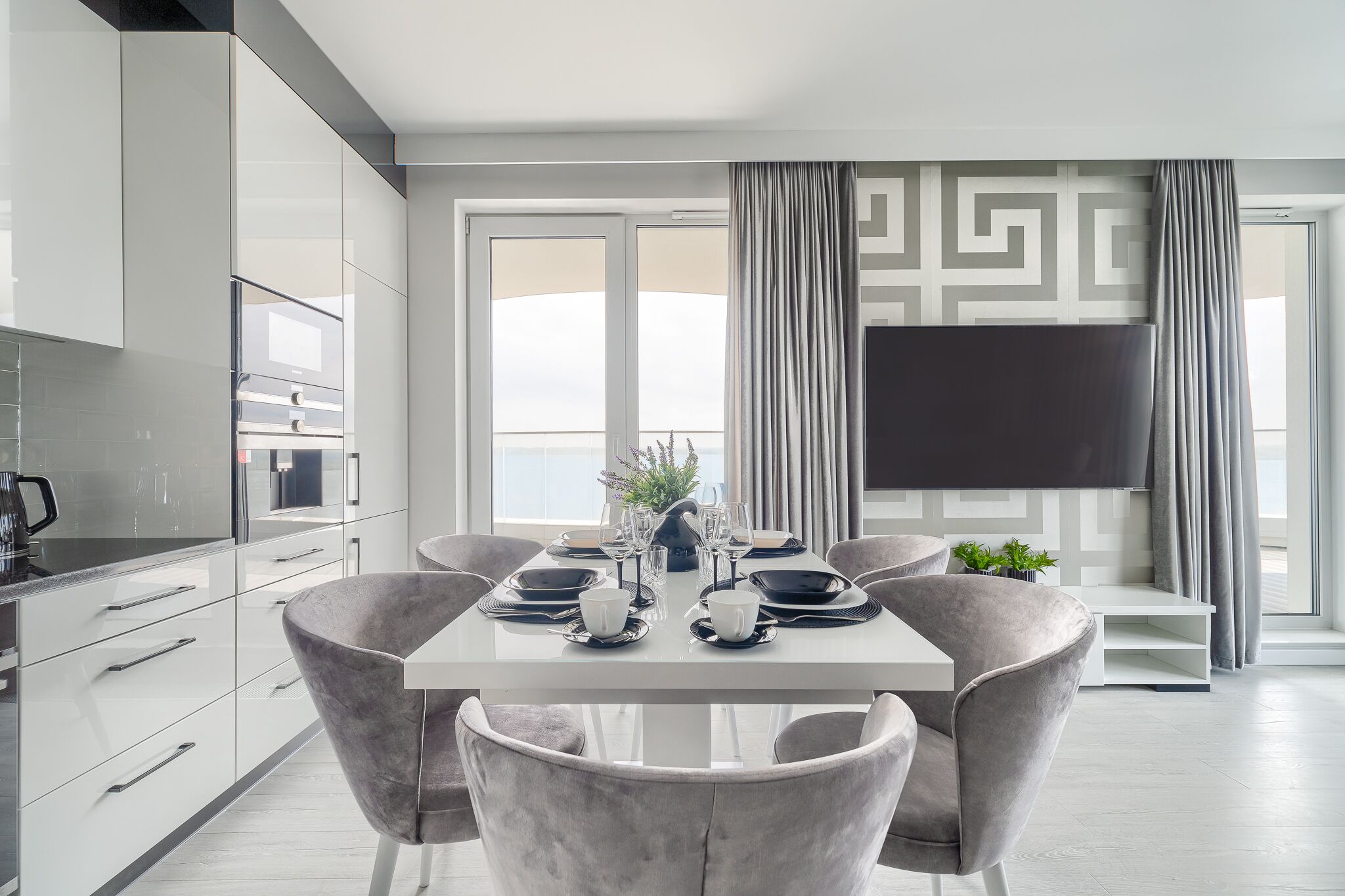 Un comedor moderno con una mesa blanca dispuesta para seis comensales, sillas grises tapizadas, un televisor de pantalla plana en la pared y vistas a la ciudad a través de amplias ventanas.