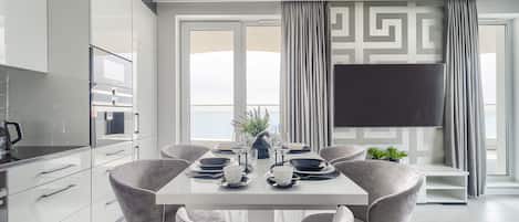 Un comedor moderno con una mesa blanca dispuesta para seis comensales, sillas grises tapizadas, un televisor de pantalla plana en la pared y vistas a la ciudad a través de amplias ventanas.