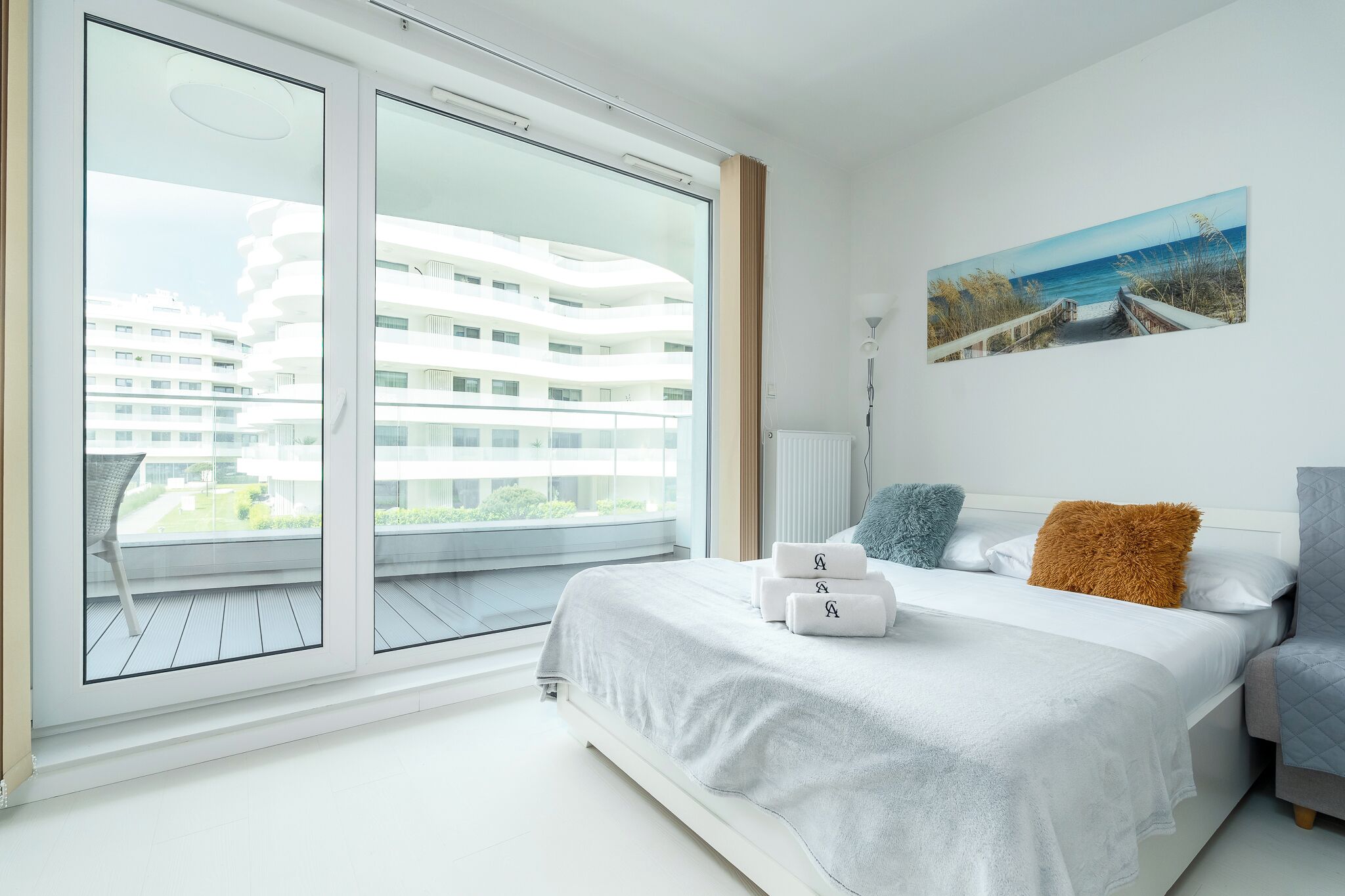 A modern hotel room with a large balcony, a bed with white bedding, and a painting of a coastal scene.