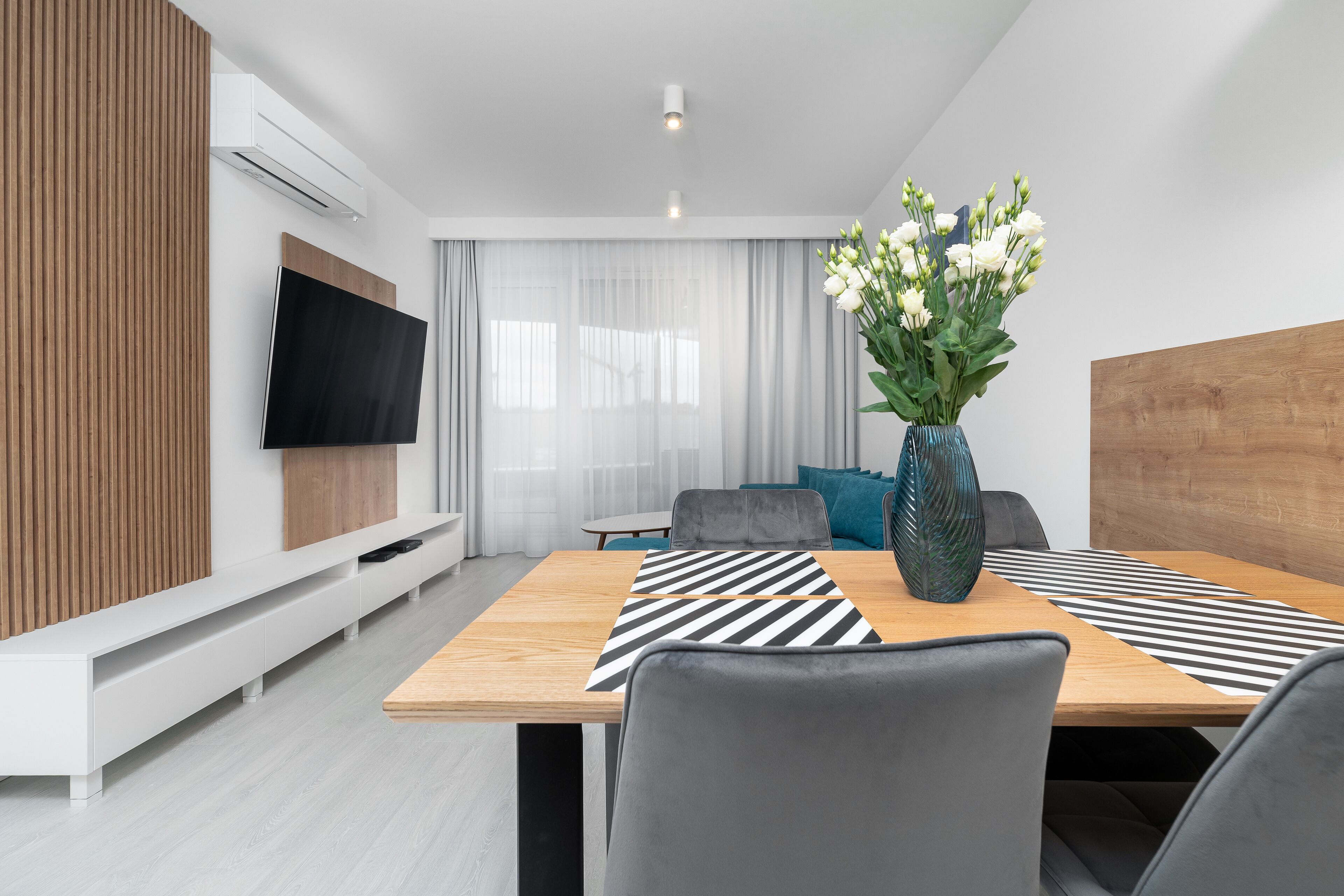 Ein modernes Wohnzimmer mit Esstisch aus Holz, grauen Stühlen, einem flachbildfernseher an der Wand, einer Holz-Akzentwand und einem großen Fenster mit transparenten Vorhängen.