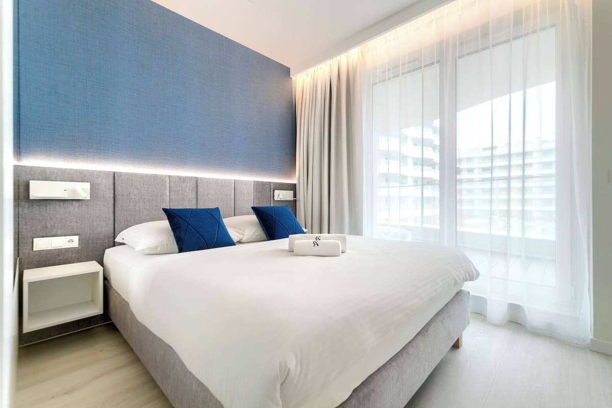Habitació d'hotel moderna amb un llit gran, dues taules de nit, un panell de control fixat a la paret i una finestra ampla amb cortines transparents.