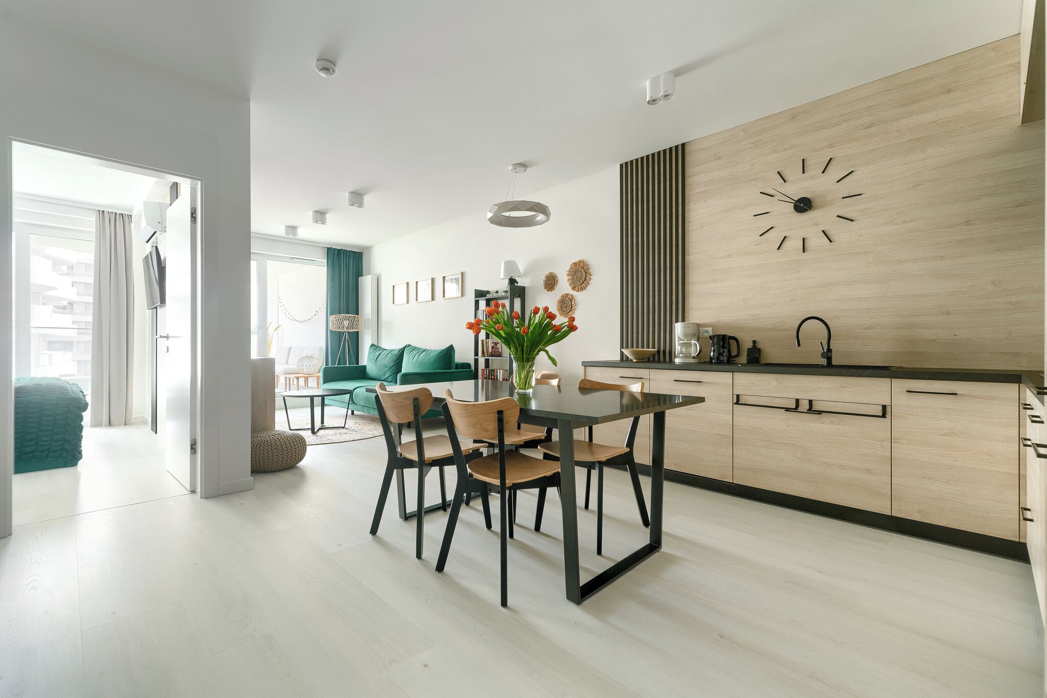 Eine moderne Küche mit einer Holz-Wand-Uhr, einem Esstisch mit Stühlen und Blick in ein Wohnzimmer mit einem grünen Sofa.