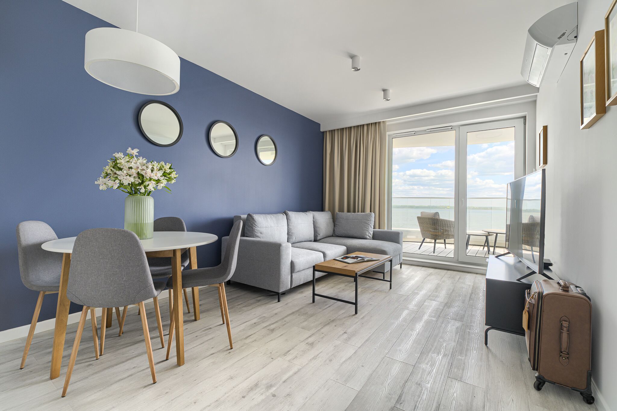 Una sala moderna con un sofá gris, una mesa redonda de comedor con sillas, una mesa de centro y un ventanal con vistas al mar.