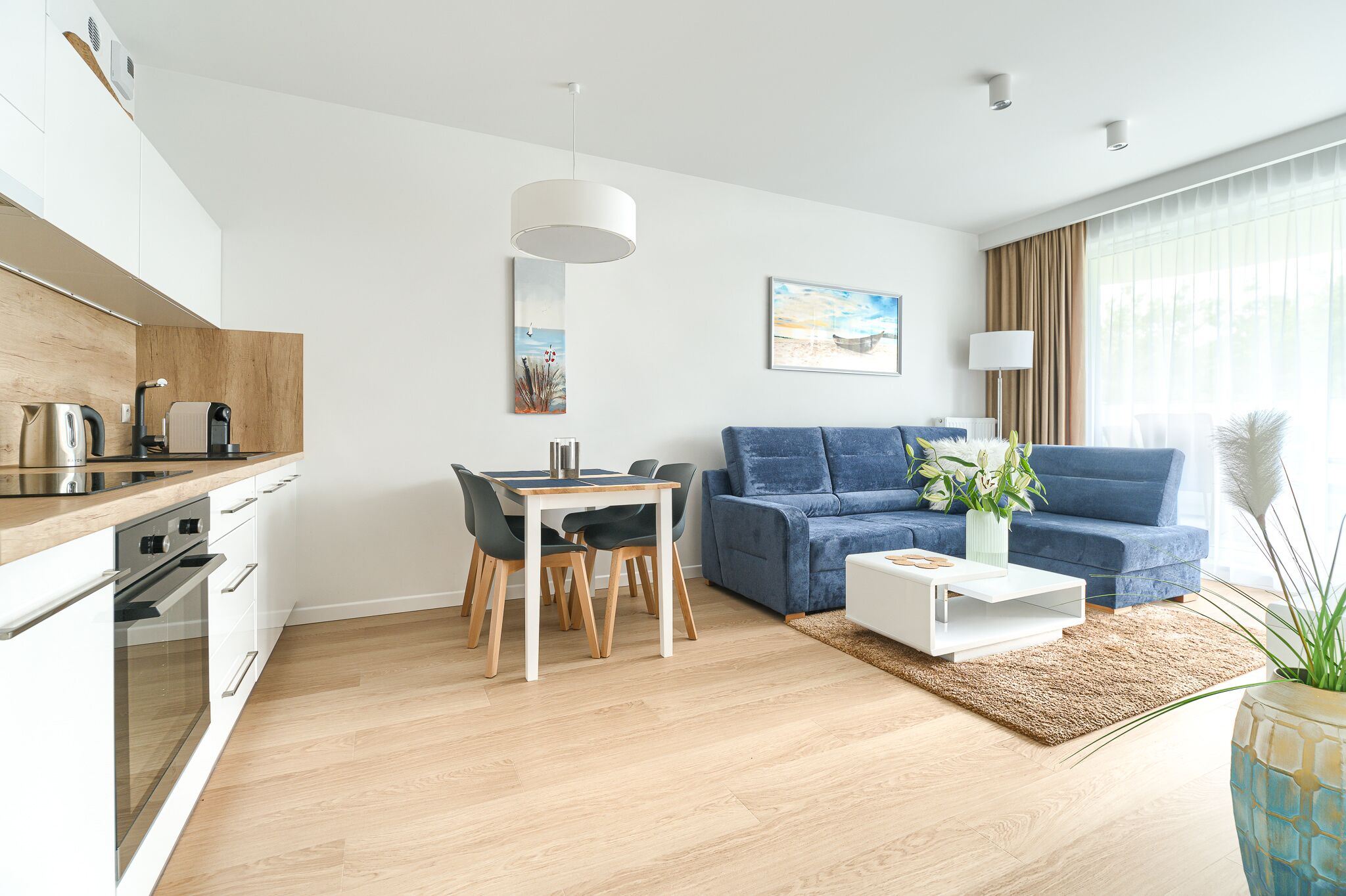 A modern living room with a blue sofa, a dining table with chairs, a white coffee table, and a kitchen area with a stove and cabinets.