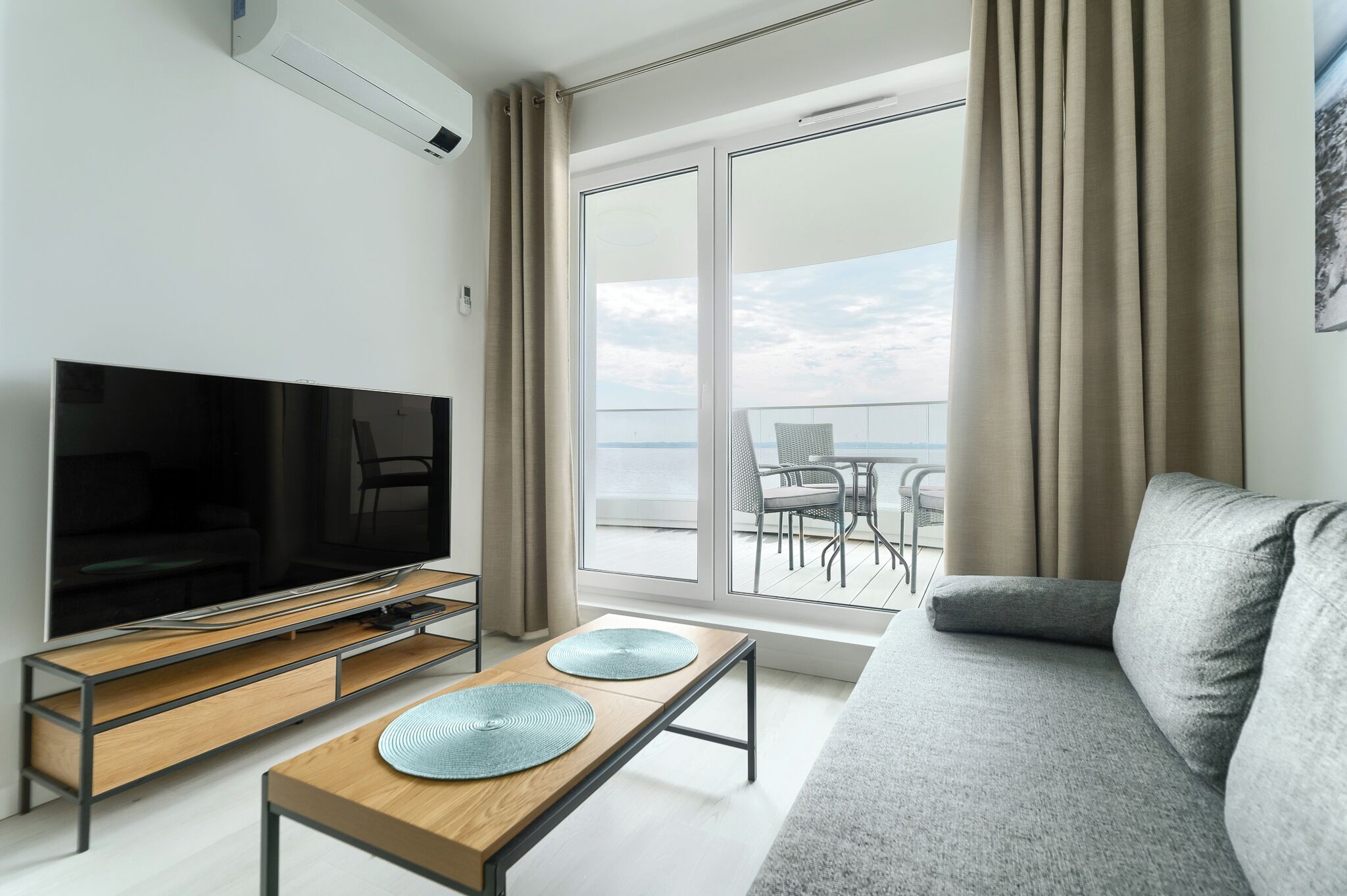 Una habitación de hotel moderna con una televisión de pantalla plana, una mesa de centro de madera con dos individuales azules, un sofá gris y un balcón con una mesa y sillas.