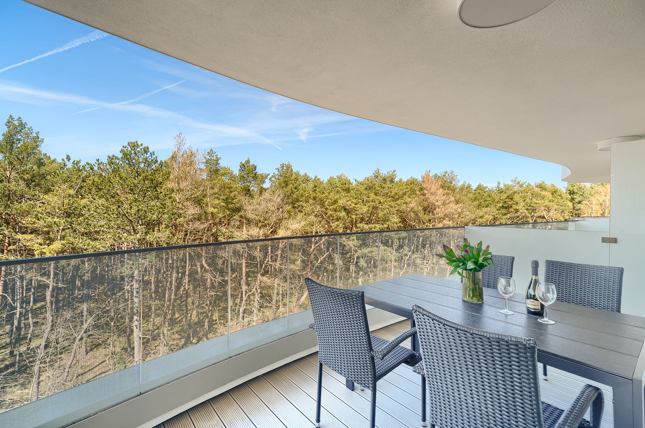 Een balkon met een tafel en stoelen, een bloemenvaas en een fles wijn.