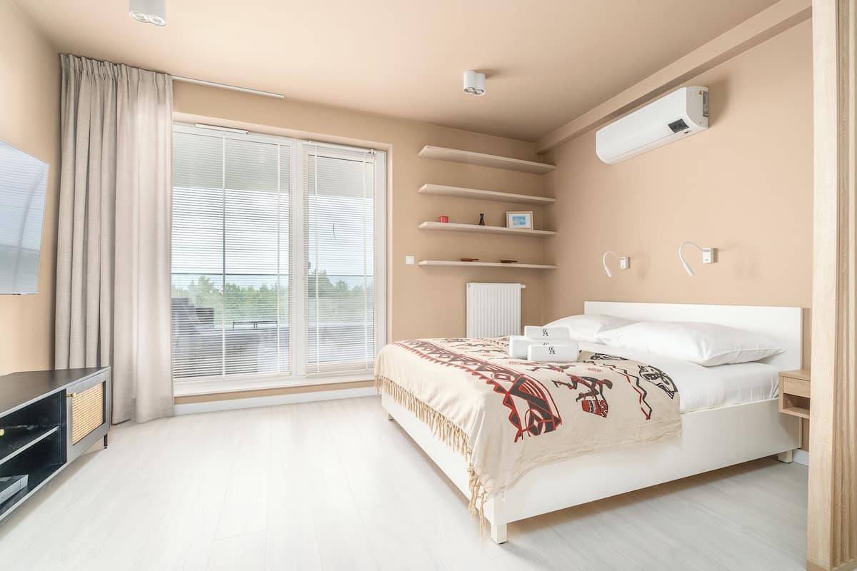 Una habitació amb un llit, una televisió, una finestra amb persianes, un ventilador de sostre i un aire condicionat fixat a la paret.