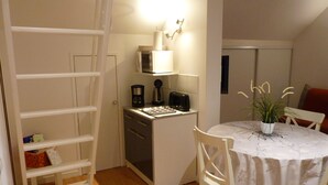 Une cuisine compacte équipée d’un four à micro-ondes, d’une machine à café et d’un grille-pain, le tout posé sur un petit plan de travail. Une chaise blanche est placée devant une petite table à manger recouverte d’une nappe blanche. Une échelle blanche est appuyée contre un mur, et une petite plante en pot est posée sur la table.